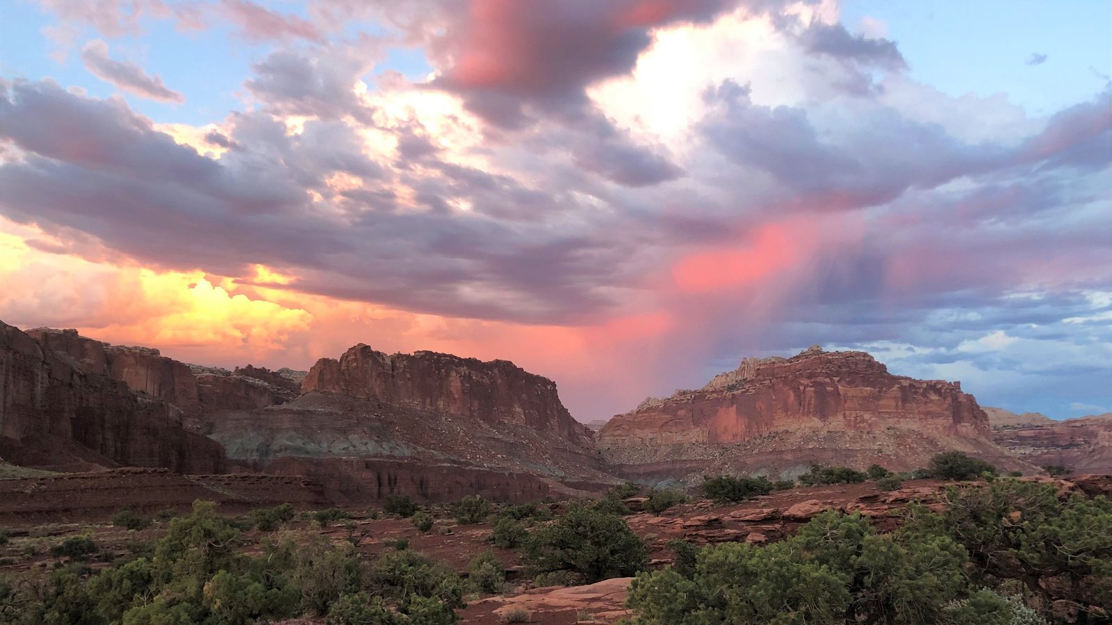 A sweeping desert canyon landscape at sunset, with layered red sandstone cliffs and buttes framing a valley, under a dramatic sky of pink- and gold-lit cloud…