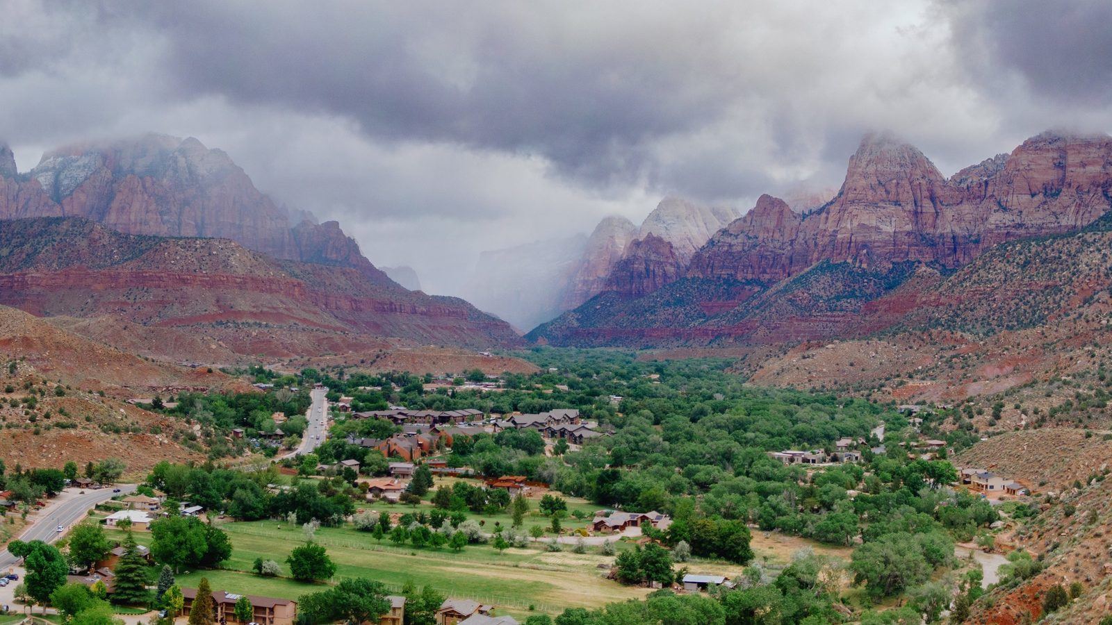 Overhead view of a small town with green trees and buildings set in a valley between tall red rock canyon walls under overcast storm clouds.