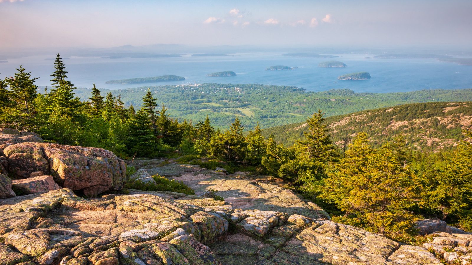 A sweeping panoramic view from the granite summit of Cadillac Mountain in Acadia National Park, Maine, overlooking forested hills, the town of Bar Harbor, an…