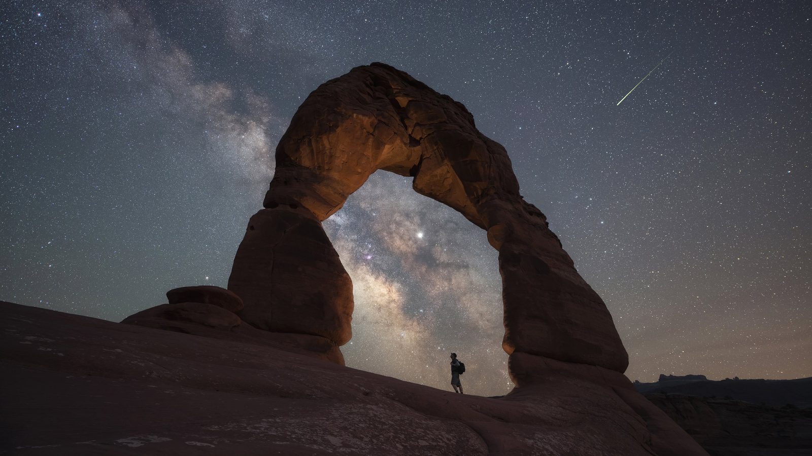 Delicate Arch in Arches National Park frames the Milky Way galaxy and a bright meteor streaking across the night sky, with a lone silhouetted hiker standing …