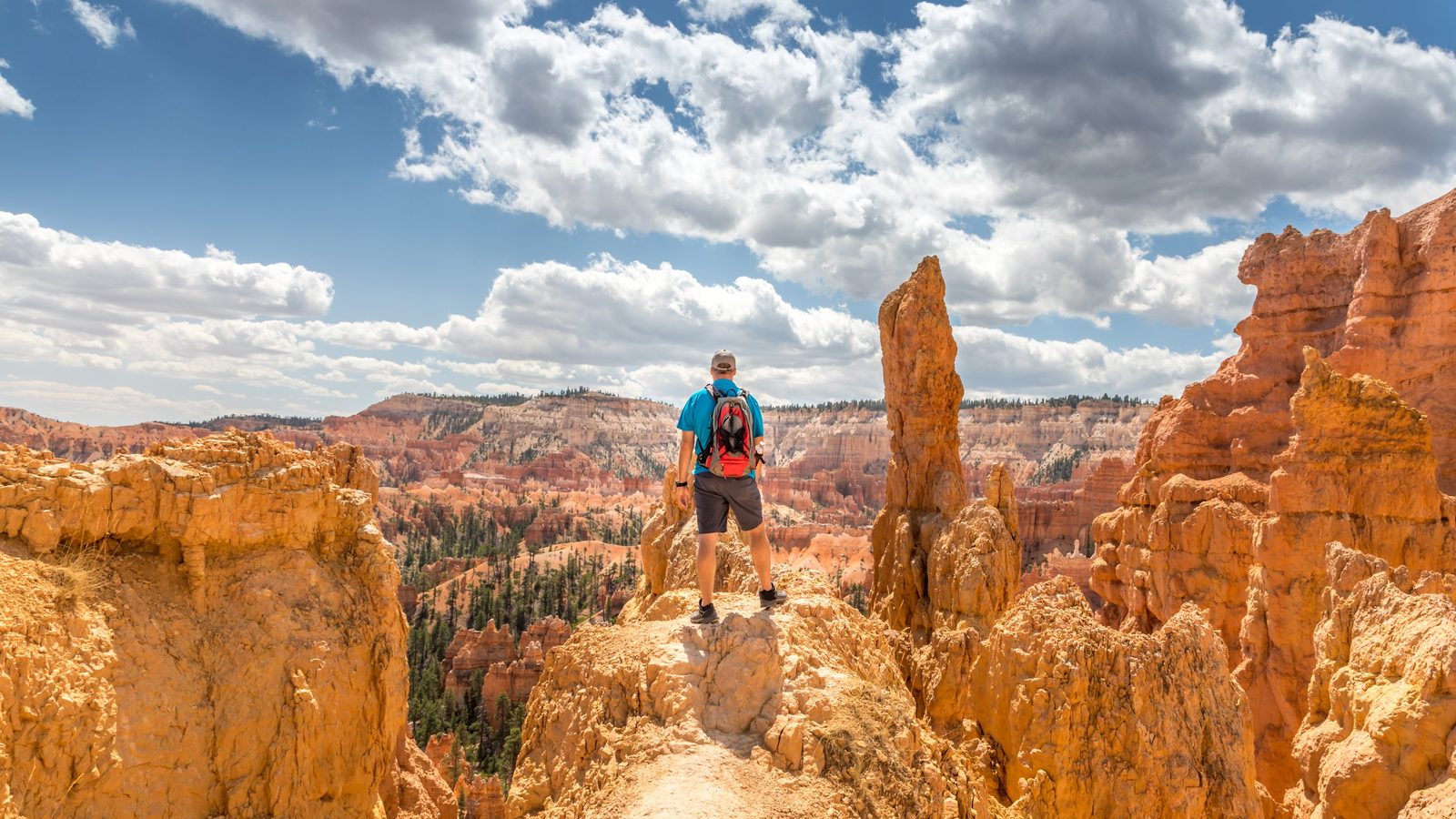 A hiker with a red backpack stands atop a rocky ridge amid towering orange-red hoodoo formations at Bryce Canyon National Park, gazing across a vast amphitheater of sculpted sandstone spires and scattered pine trees under a dramatic partly-cloudy sky.