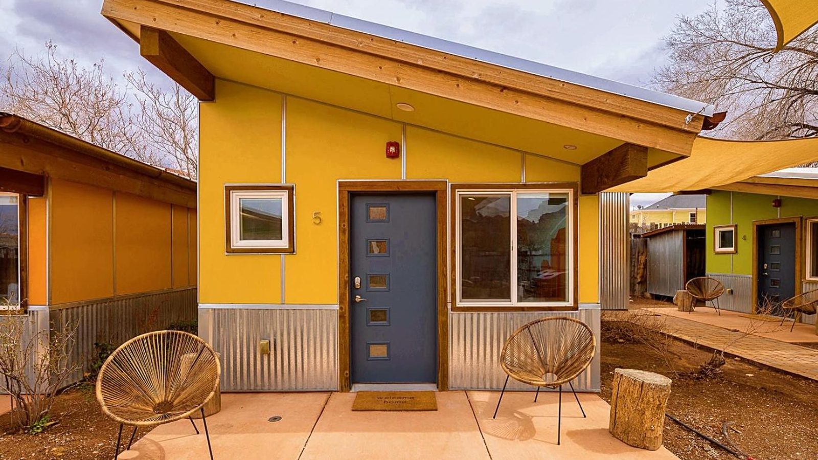 A brightly painted yellow vacation cabin or motel unit numbered '5' with a blue mid-century modern door, corrugated metal siding, and two woven Acapulco chai…