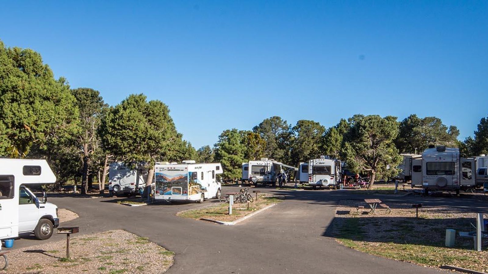 An RV campground at what appears to be the Grand Canyon South Rim, with multiple motorhomes and travel trailers parked along a paved loop road surrounded by …