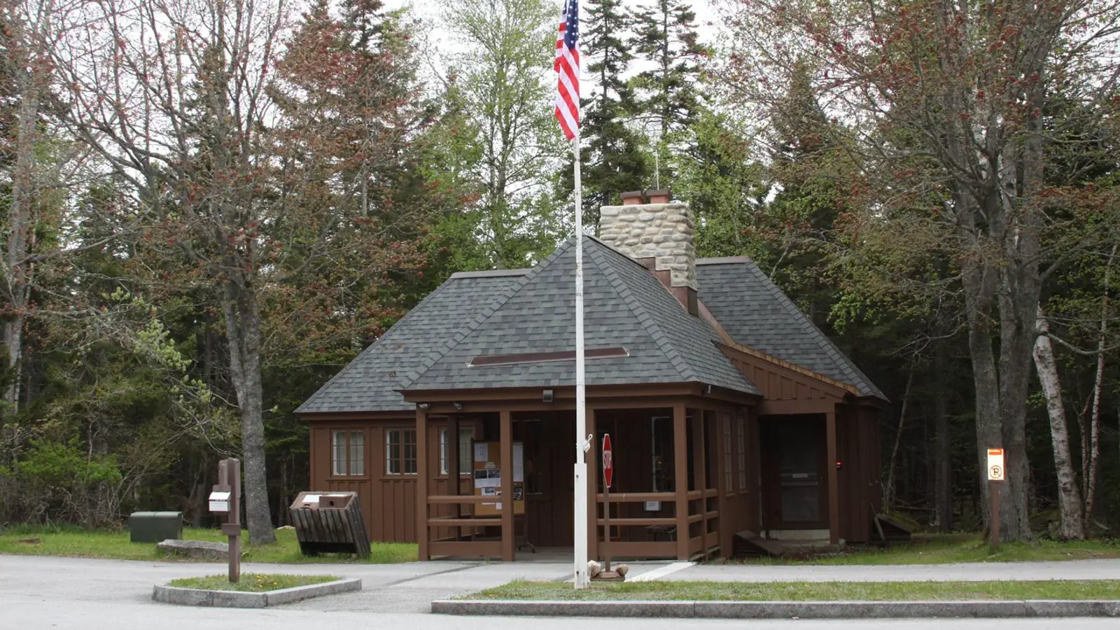 A small brown wooden ranger station or visitor contact building with a dark shingle hip roof and stone chimney, surrounded by mixed deciduous and evergreen t…