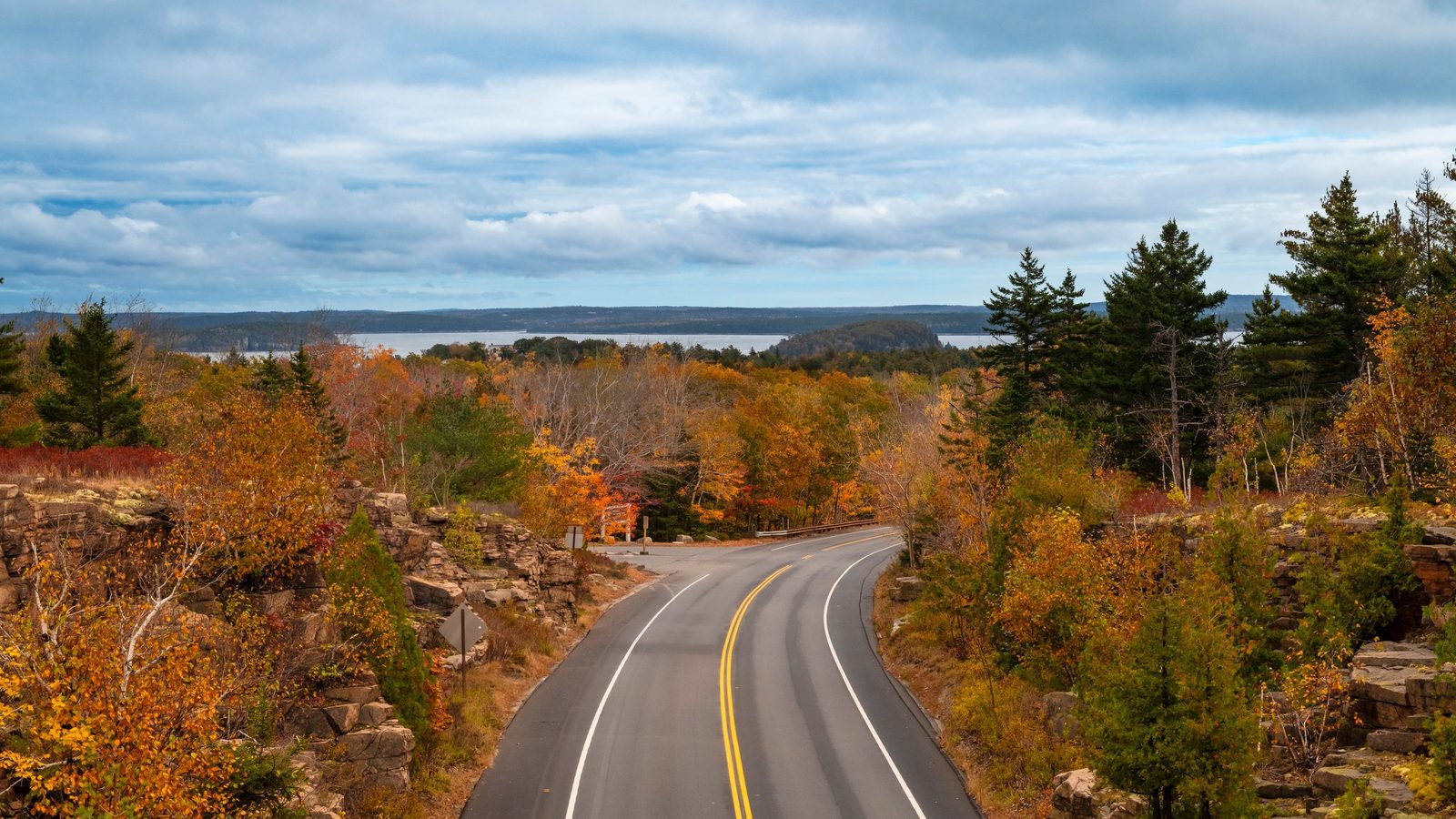 A winding two-lane road curves through a vibrant autumn forest in Acadia National Park, with fiery orange, red, and yellow foliage flanking rocky outcropping…