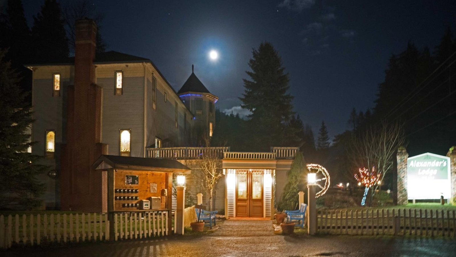 Alexander's Lodge, a Victorian-style inn, is photographed at night under a full moon with warm interior lighting glowing from its windows and entry, holiday …