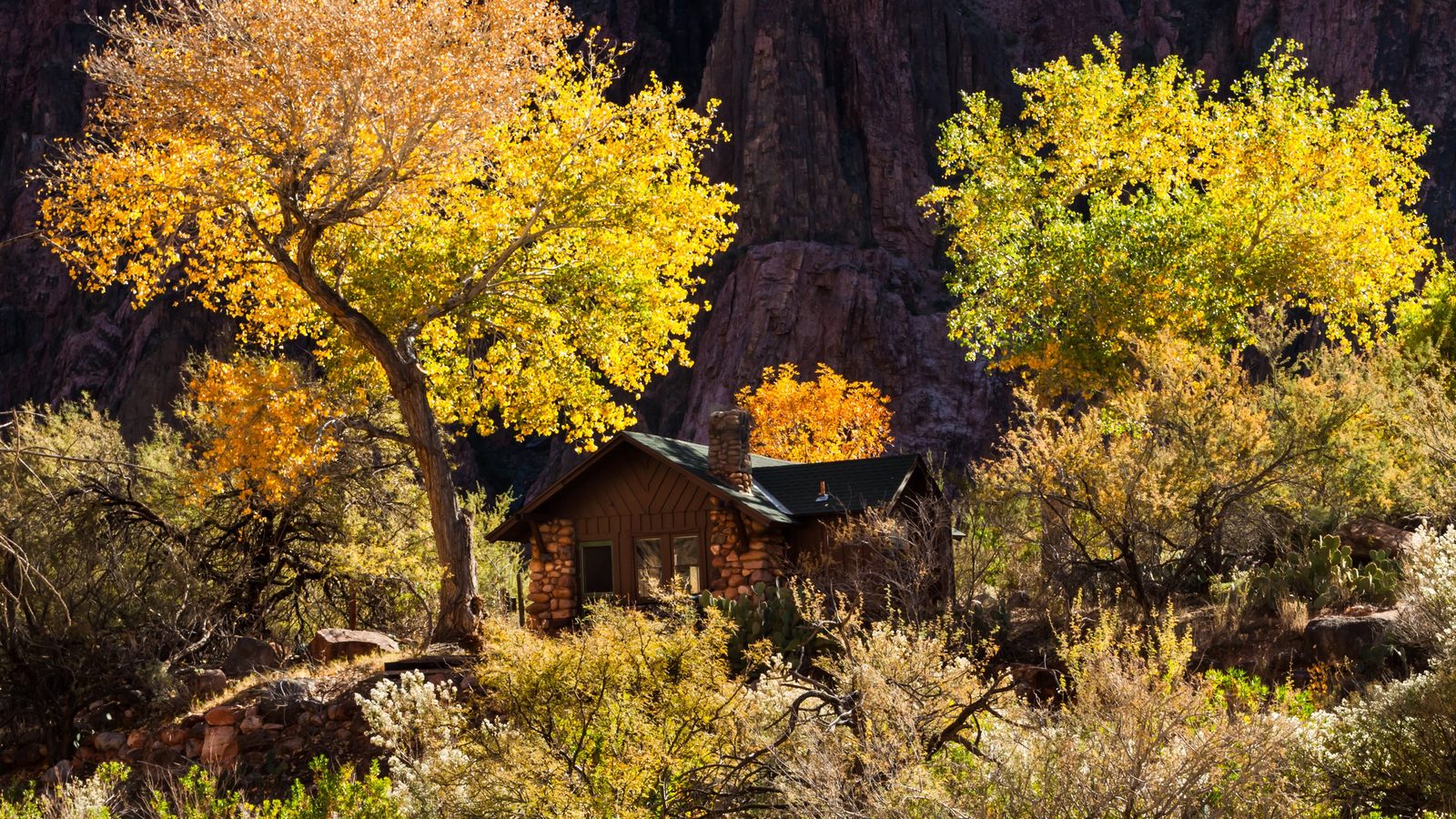 A rustic stone cabin nestled among vibrant golden-yellow cottonwood trees in full autumn color, set against dramatic dark red canyon walls that create a stri…