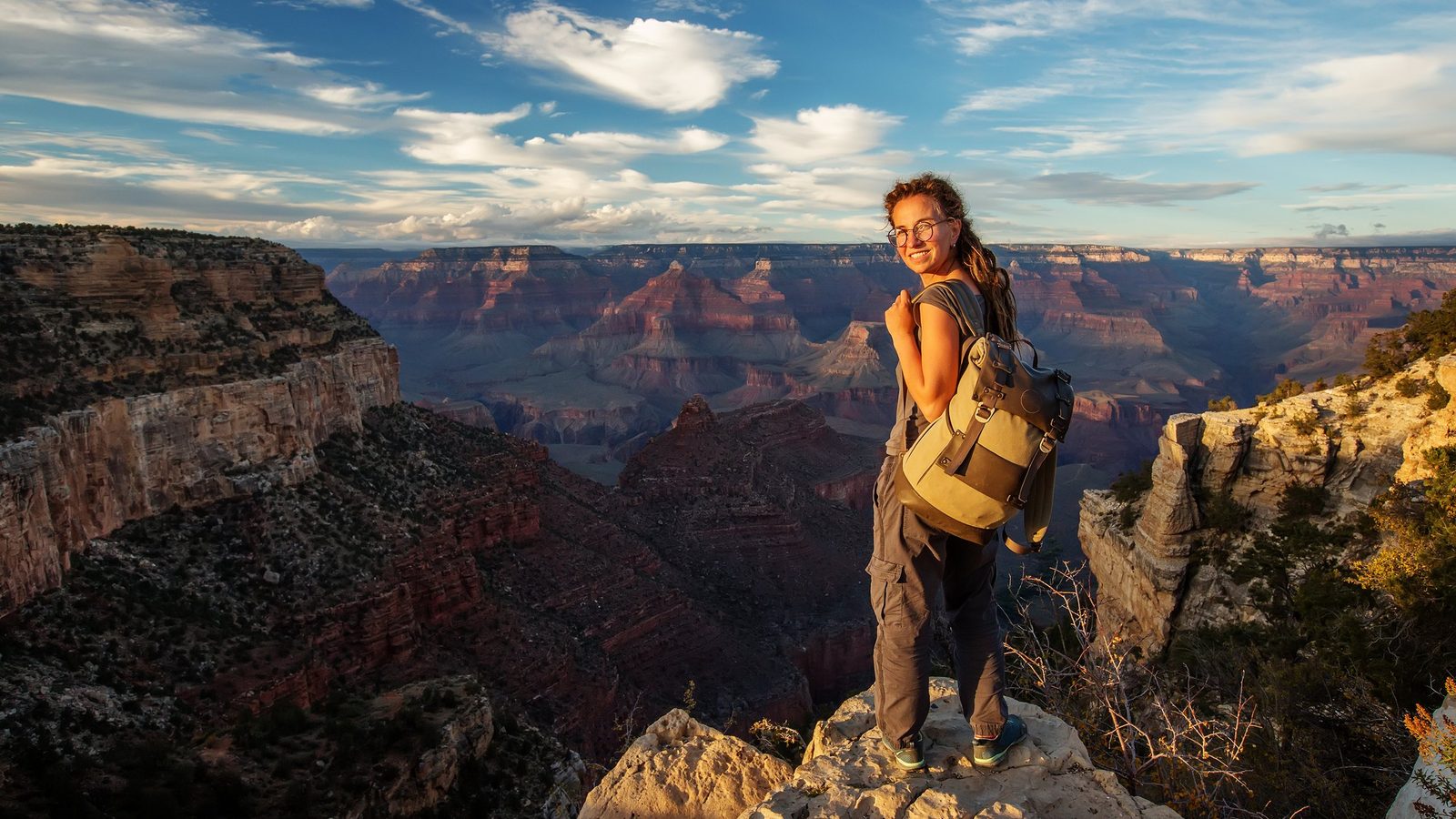 A smiling female hiker with a backpack stands on a rocky outcrop at the Grand Canyon's South Rim, with vast layered red-rock canyon formations stretching int…