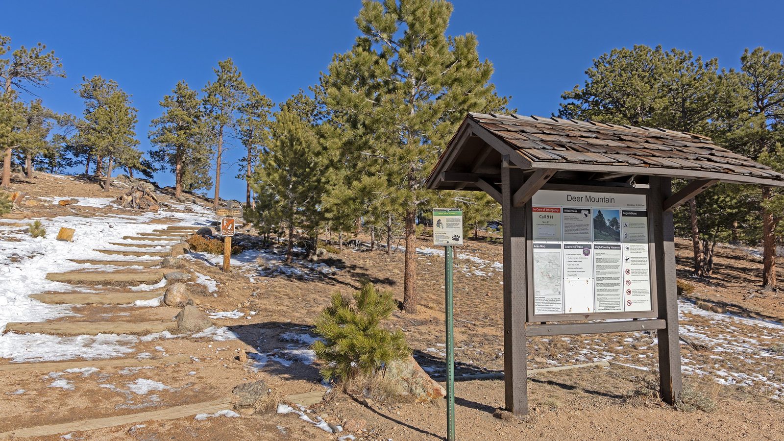 The trailhead kiosk for Deer Mountain in Rocky Mountain National Park stands beside a sandy, partially snow-dusted path with wooden step reinforcements leadi…