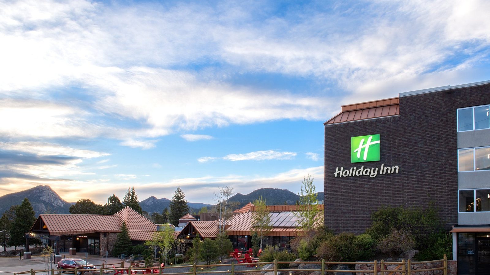 A Holiday Inn hotel exterior is shown at dusk with a rustic mountain-lodge aesthetic, featuring copper-roofed buildings, wood fencing, and red Adirondack cha…