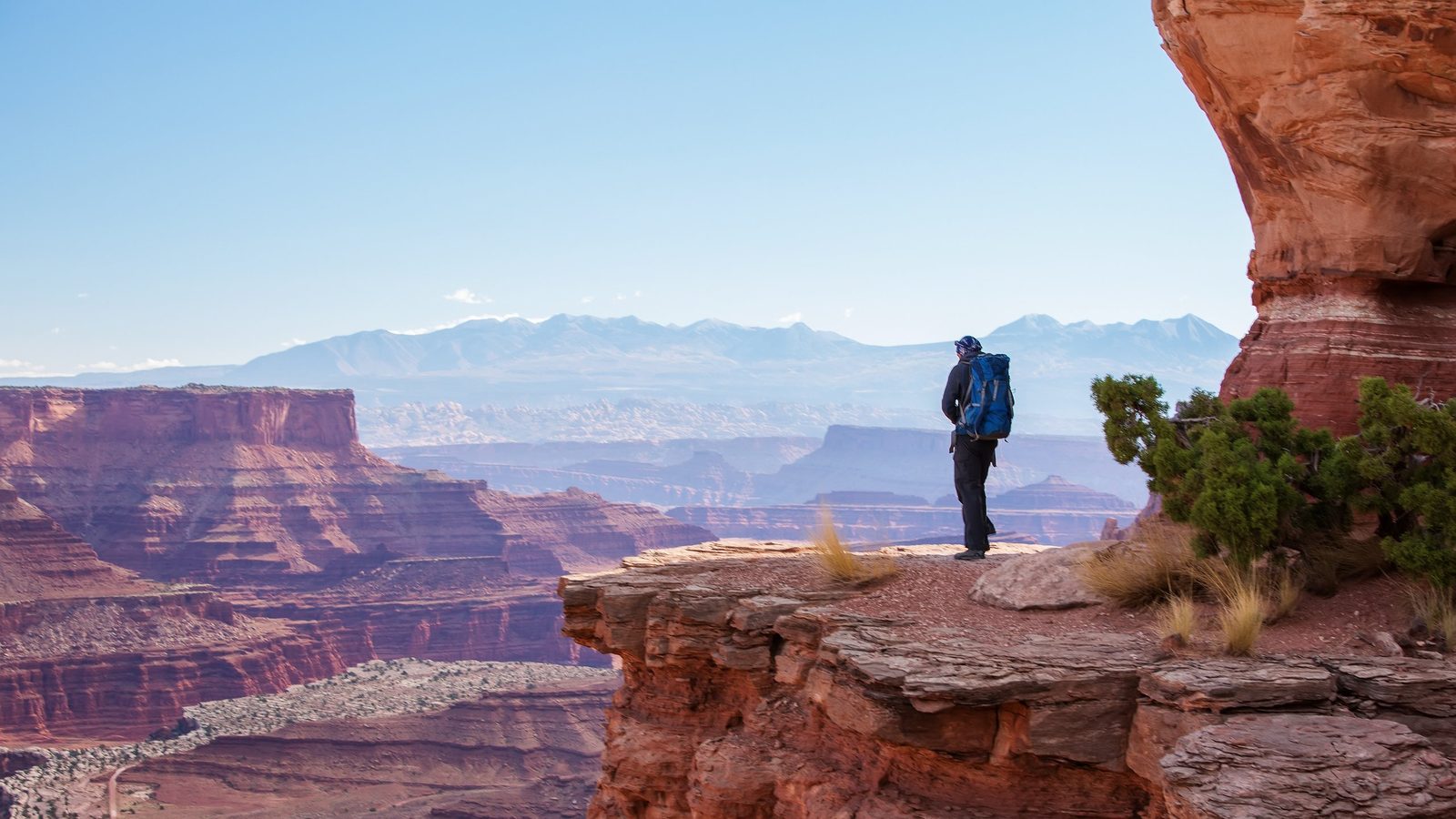 A lone hiker with a blue backpack stands on the edge of a dramatic red sandstone cliff overlooking a vast canyon landscape at Canyonlands National Park, with…