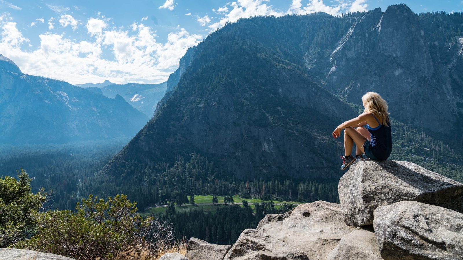 A lone hiker sits on a granite outcrop overlooking the sweeping Yosemite Valley, with towering granite cliffs, dense conifer forests, and a lush valley floor…
