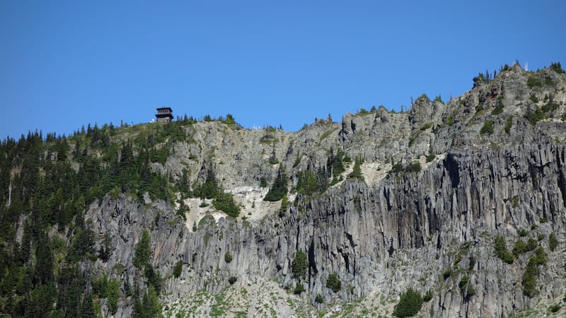 A sheer basalt cliff face rises steeply above a rocky talus slope, with scattered subalpine fir trees clinging to ledges and a small fire lookout tower perch…