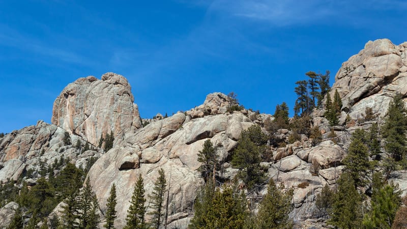 Large granite rock outcroppings tower above a hillside forest of green conifers against a clear blue sky.