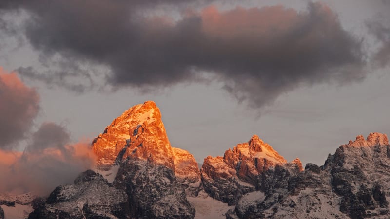 The jagged snow-dusted peaks of the Grand Teton range are bathed in warm alpenglow at sunrise or sunset, with the tallest summit glowing deep orange against …