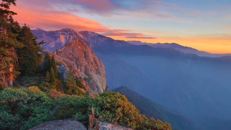 A dramatic sunset view from Moro Rock in Sequoia National Park, showcasing a massive granite dome flanked by conifers and manzanita shrubs, with layered mist…