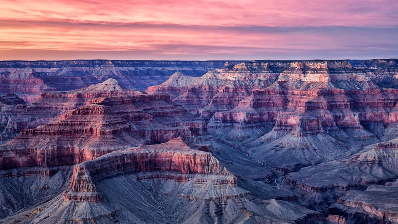 Aerial-perspective view of the Grand Canyon at twilight, showing layers of red and gray sandstone buttes receding toward the North Rim under a pink and magen…