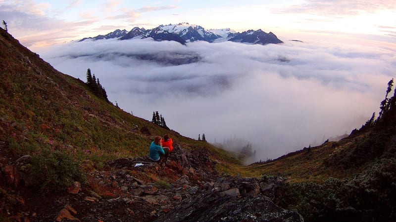 Two hikers sit on a rocky ridge above a sea of clouds, gazing toward a snow-capped mountain range emerging above the cloud layer at twilight.