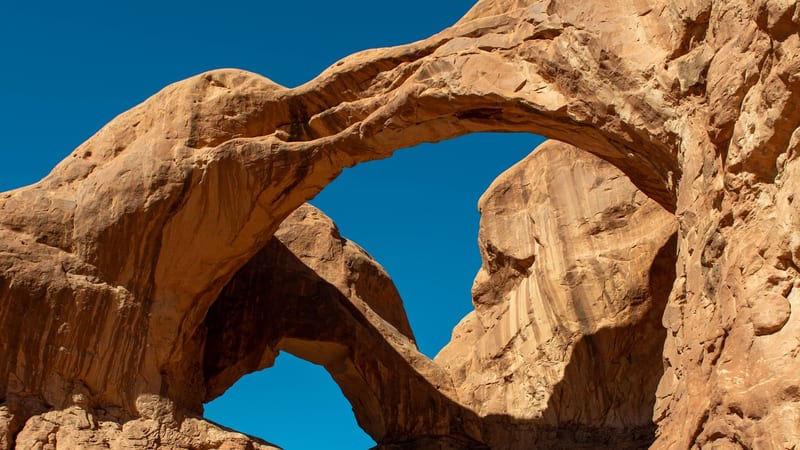 Double Arch, a massive pair of natural sandstone arches sharing a common wall, rises against a clear deep-blue sky in Arches National Park; warm midday sunli…