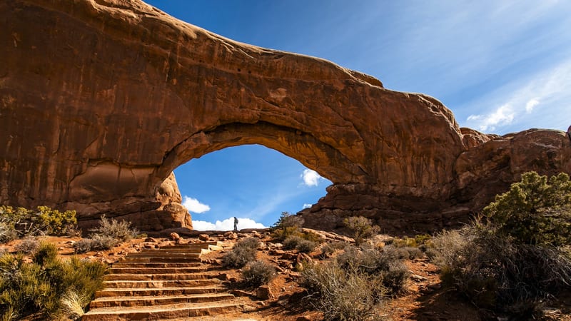 A massive red sandstone arch frames a vivid blue sky at Arches National Park, with stone staircase steps leading up through desert scrub toward a lone hiker …