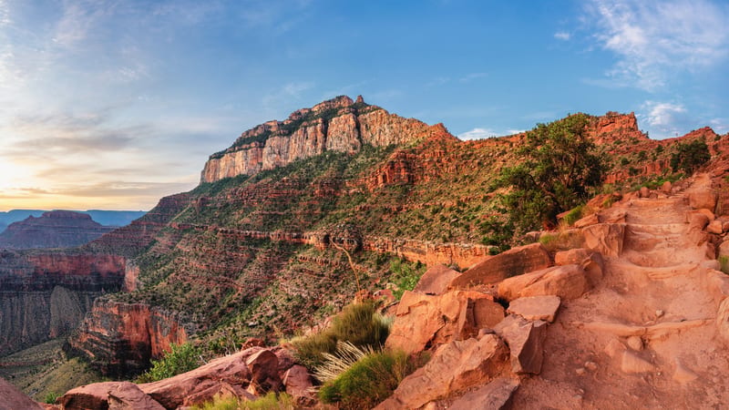 A stone-stepped hiking trail winds down the South Rim of the Grand Canyon past red sandstone rocks and green shrubs, with a tall pale limestone butte and lay…
