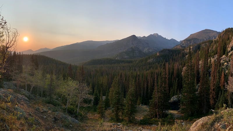 A wide panoramic view of a forested mountain valley at sunrise or sunset in Rocky Mountain National Park, with a low sun casting warm orange hues through haz…
