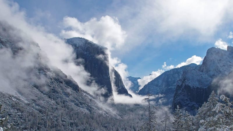 El Capitan, the massive granite monolith of Yosemite Valley, rises vertically through low mist and clouds against a deep blue sky, with its pale face reflect…