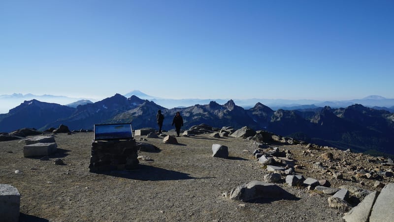 A high-alpine rocky summit plateau at Mount Rainier National Park, with two hikers near an interpretive sign and a sweeping panorama of the Tatoosh Range and…