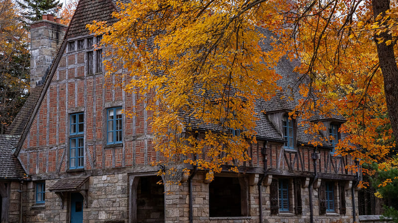 A Tudor Revival or English cottage-style stone and half-timbered manor house is surrounded by autumn foliage in brilliant shades of gold, orange, and red.