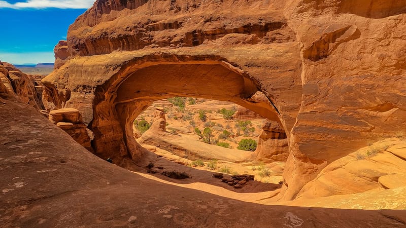 A large natural sandstone arch frames a sweeping desert canyon view beyond, with warm orange-red rock layers stacked above and vivid blue sky with wispy clou…