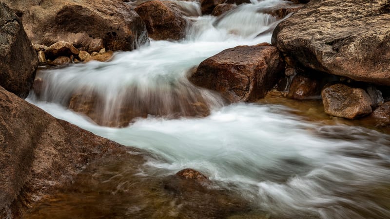 A fast-moving mountain stream flows over and around large reddish-brown boulders, with long-exposure blur giving the white water a smooth, silky appearance.