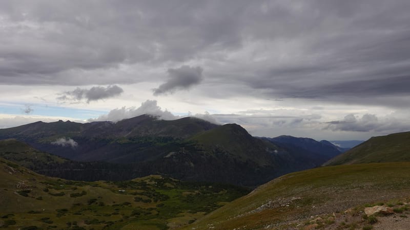 A high-elevation alpine landscape shows rolling tundra in the foreground giving way to a broad mountain valley and a series of dark, forested ridgelines unde…