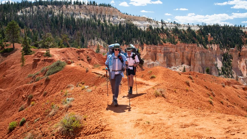 Two backpackers with trekking poles and large packs hike along a red-dirt trail through Bryce Canyon's interior, surrounded by vivid orange-red hoodoos and e…