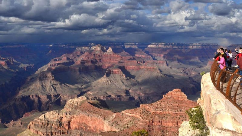 A panoramic view of the Grand Canyon's South Rim from a railed overlook, where layered red and orange sandstone buttes and mesas stretch toward the horizon u…