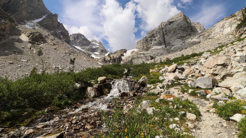 A rushing glacial stream cascades over boulders through a high alpine valley in the Teton Range, framed by rugged gray granite peaks with remnant snow patche…