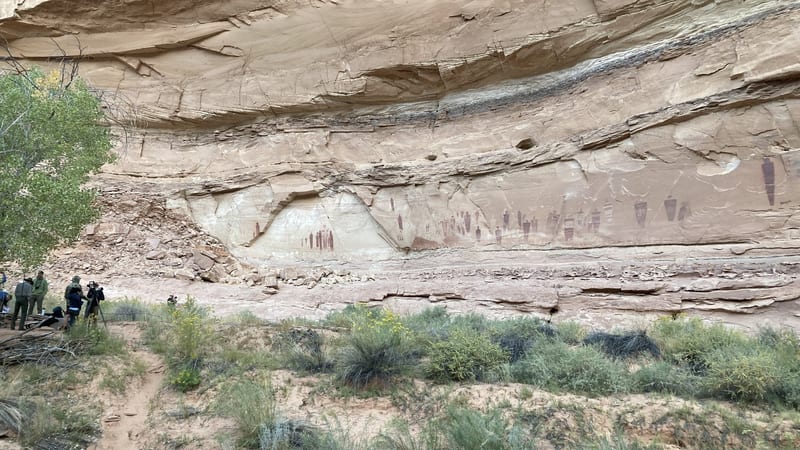 A massive sandstone canyon wall displays a long panel of ancient Native American pictographs painted in dark reddish-brown, with researchers and camera equip…