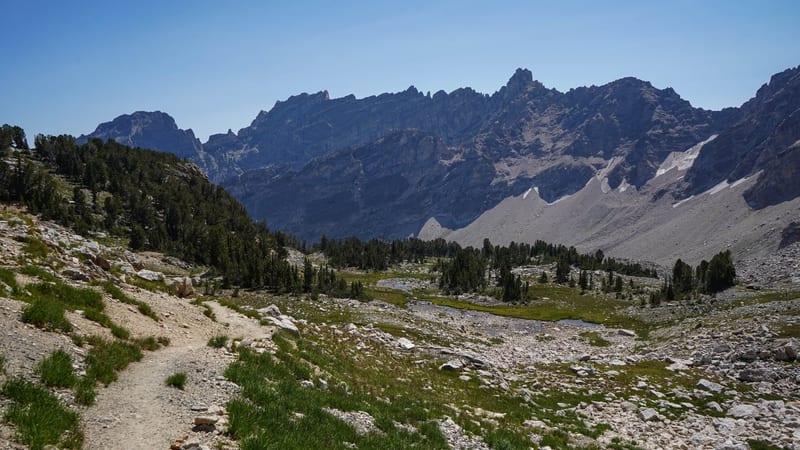 A rocky alpine valley trail winds through a high-elevation Sierra Nevada landscape, with scattered conifers, a small meadow stream, and jagged granite peaks …
