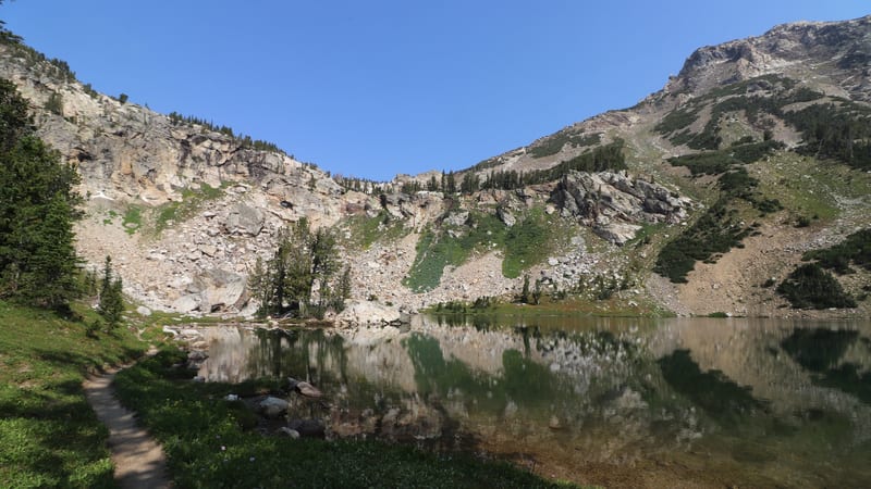A calm alpine lake sits in a glacially carved cirque, its still surface perfectly mirroring the surrounding rocky slopes, talus fields, and scattered conifer…
