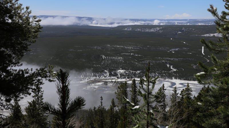 A high-elevation winter overlook in Yellowstone National Park shows a snow-dusted river valley with a meandering partially frozen river, dense conifer forest…