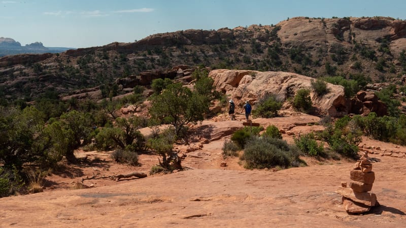A wide sandstone slickrock trail winds through sparse juniper shrubs in a red-rock desert landscape, with two hikers in blue jackets walking toward a rocky r…