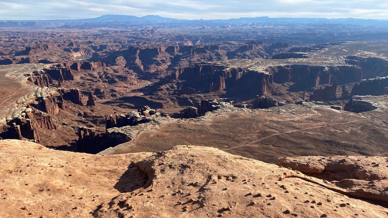 A sweeping aerial-perspective view from a sandstone mesa rim over the vast canyon labyrinth of Canyonlands National Park, with warm reddish-orange rock in th…