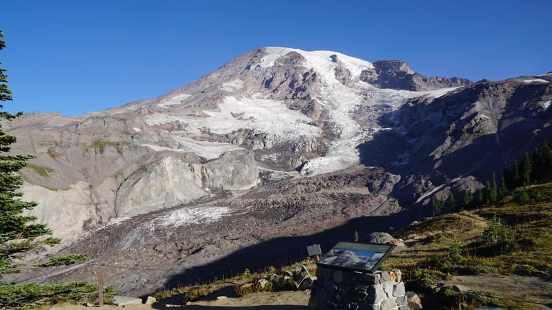A receding glacier on the flanks of Mount Rainier fills the frame, revealing extensive bare rock and moraine where ice has retreated, under a clear blue late…