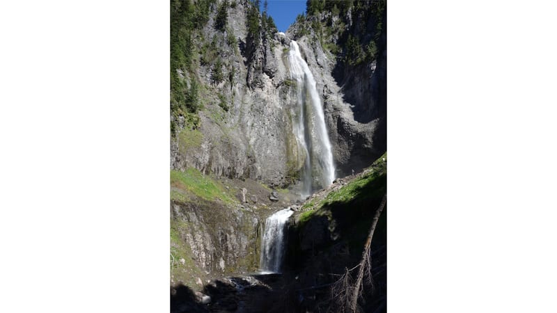 A tall multi-tiered waterfall cascades down a sheer volcanic rock cliff face framed by evergreen trees, with bright midday sunlight illuminating the white wa…