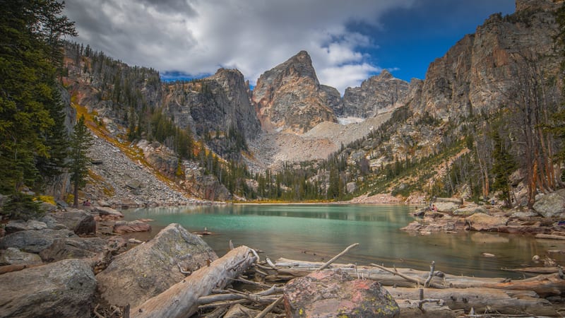 A calm alpine lake reflects the jagged, snow-streaked peaks of the Teton Range under a clear blue sky, framed by rocky canyon walls and a band of conifer tre…