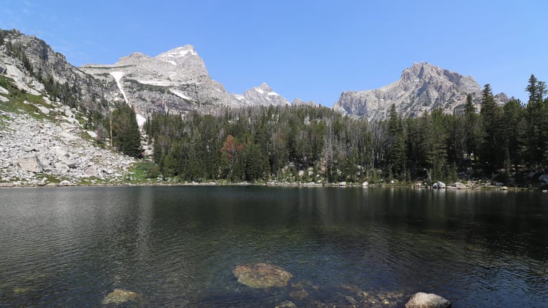 A clear alpine lake with a rocky, transparent-bottomed foreshore reflects a dense conifer forest and jagged granite peaks dusted with snow under a bright blu…