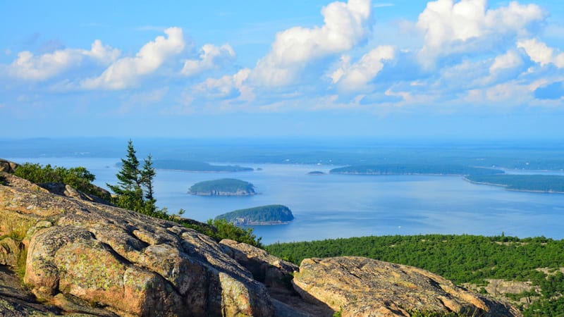 View from a granite mountain summit in Acadia National Park looking east over Frenchman Bay, with green forested islands dotting the calm blue water and whit…