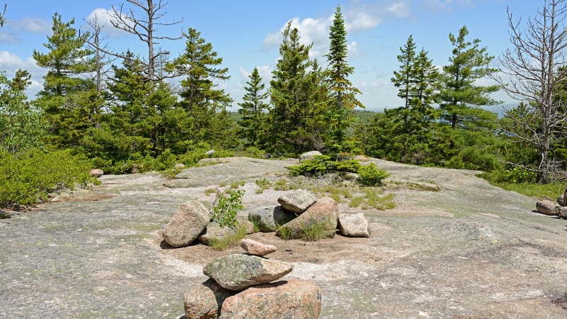 Flat granite summit ledge with scattered boulders and tufts of grass, surrounded by spruce and pine trees under a blue sky with white clouds.