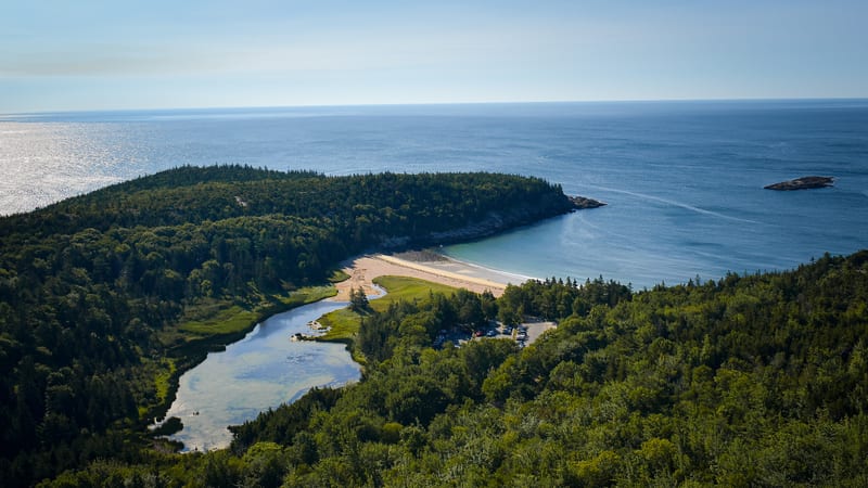 Aerial view of a crescent-shaped sandy beach bordered by dark green conifer forest, with a freshwater stream and lagoon in the foreground and the open Atlant…
