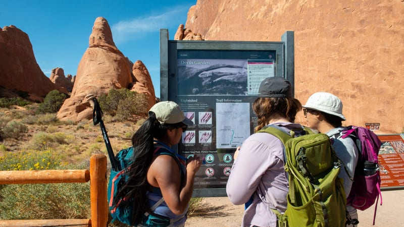 Three hikers study a Devils Garden trail information sign at Arches National Park, surrounded by red sandstone formations under a blue sky.