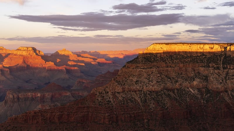 A panoramic view across the Grand Canyon at sunset, with layered red and orange sandstone buttes and mesas lit by warm golden light receding into a hazy blue…
